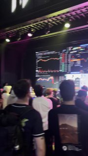 Зрители следят за трансляцией трейдов: толпа перед большим экраном с графиками BTC/USDT и интерфейсом биржевых сделок, размыто.