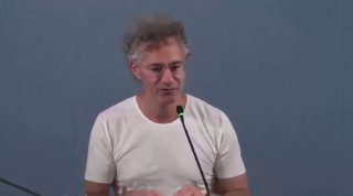Кадр из видео: человек в светлой футболке стоит у микрофона на фоне серой стены; иллюстрация к материалу о Palantir, личность на фото не подписывается.