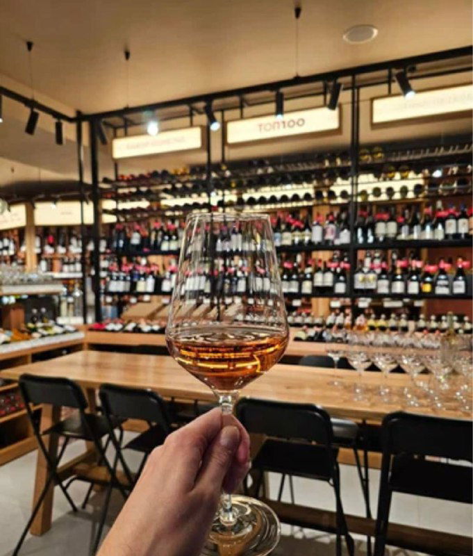 Бокал вина в руке на фоне винного магазина с рядами бутылок и барной стойкой — атмосфера винотеки перед началом дегустации.