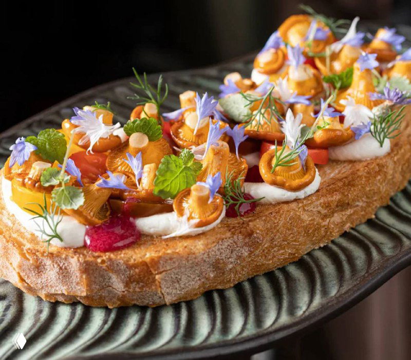 Брускетта с обжаренными лисичками, страчателлой и съедобными цветками — элегантная закуска на хрустящем хлебе с травами.