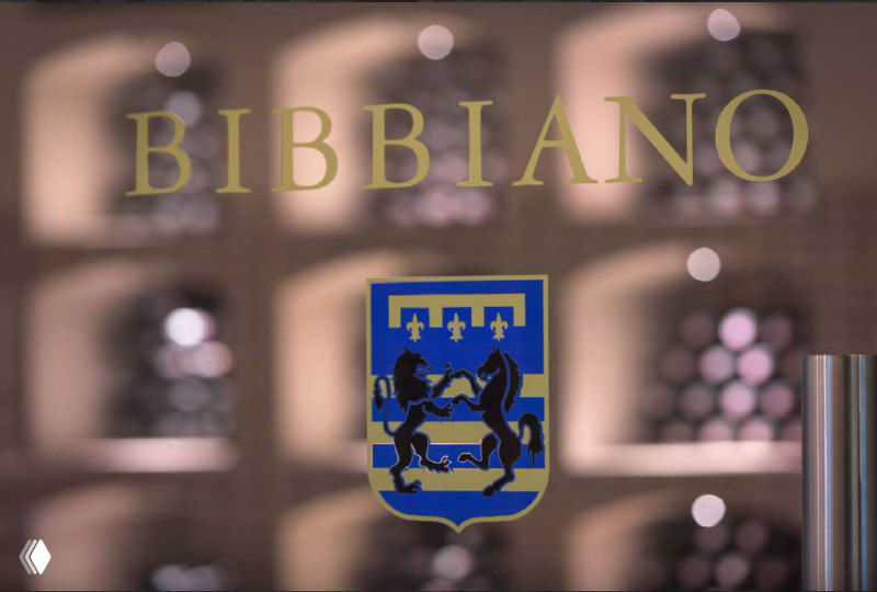 Оформление с надписью BIBBIANO и гербом винодельни Tenuta di Bibbiano на размытом фоне интерьера и стеллажей с бутылками.