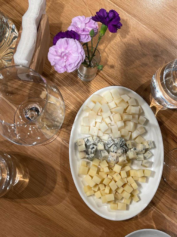 Крупный план сырной тарелки и бокалов на столе дегустации: мелко нарезанные кубики сыра, цветы в вазе и отражения стекла в кадре.