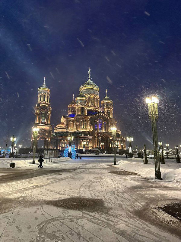 Ночной вид храма ВС России со снежной площадкой: подсветка здания и люди прогуливаются по снежным аллеям возле храма.