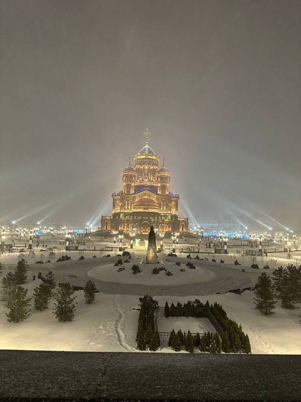 Вид храма ВС России в снегопад: освещённый фасад, заснеженные клумбы и ровная симметрия подхода к входу.