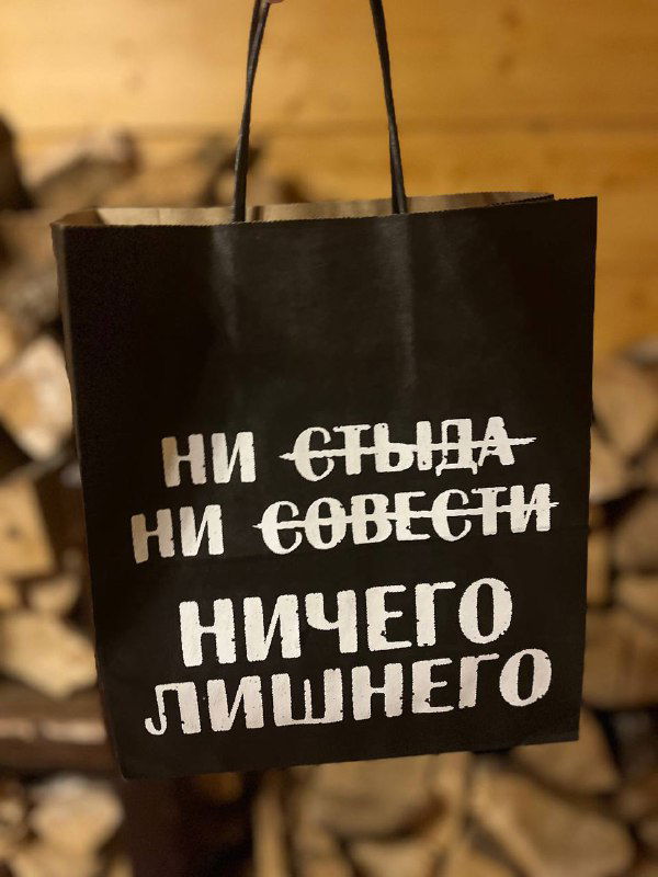 Черный подарочный пакет с белой надписью «Ни стыда ни совести ничего лишнего», подвешен на фоне дров и деревянной стены.
