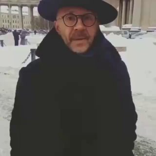 Кадр видео: мужчина в тёмной шляпе и очках на заснеженной городской площади, близкий план, уличная сцена с прохожими за спиной.