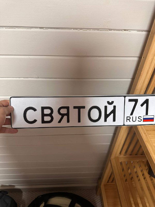 Фотография автомобильного номера с надписью «СВЯТОЙ 71» и российским флажком, лежащего на деревянной поверхности внутри помещения.