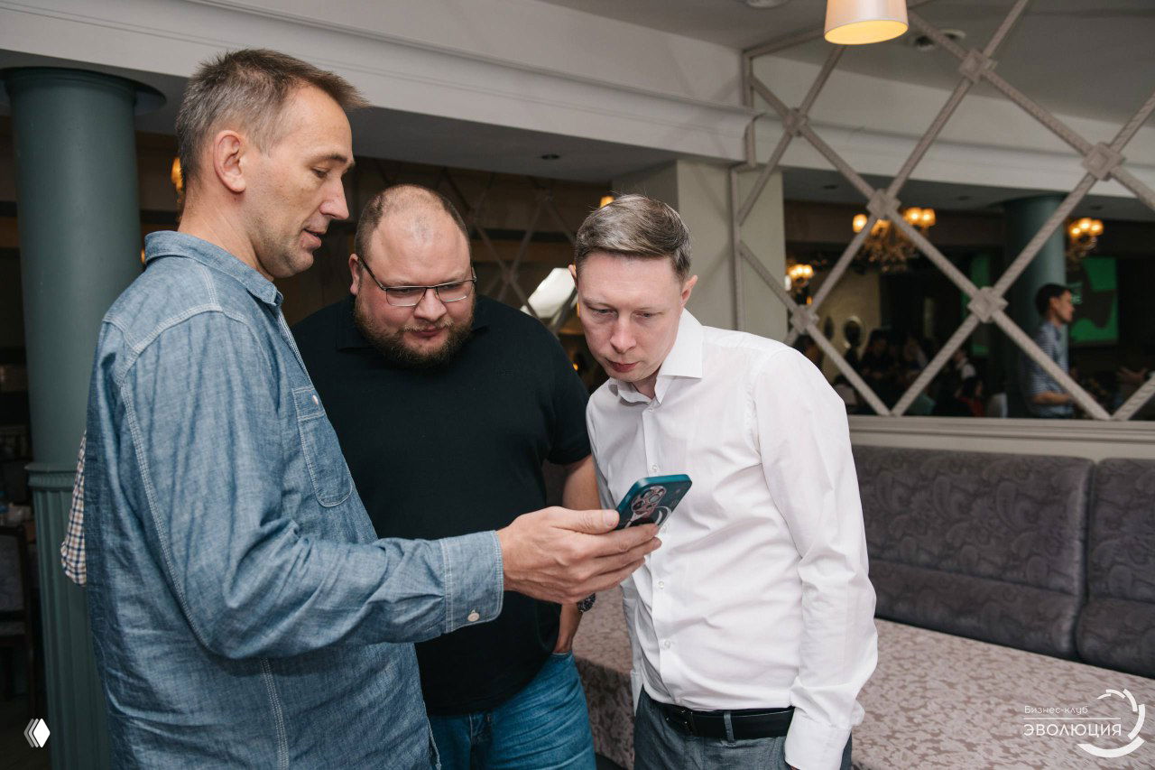 Три участника конференции внимательно смотрят в экран смартфона в перерыве, неформальное общение между предпринимателями.