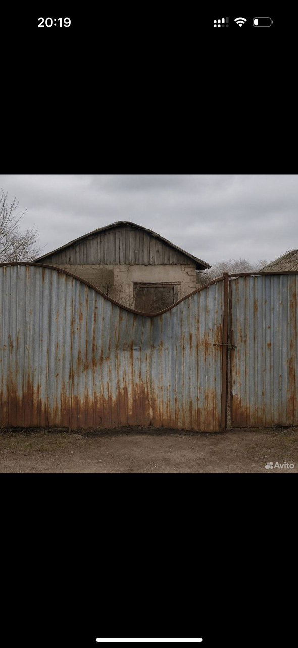 Фото из объявления на Авито: волнообразный ржавый забор с воротами и старыми постройками на заднем плане под пасмурным небом, индустриальный антураж.