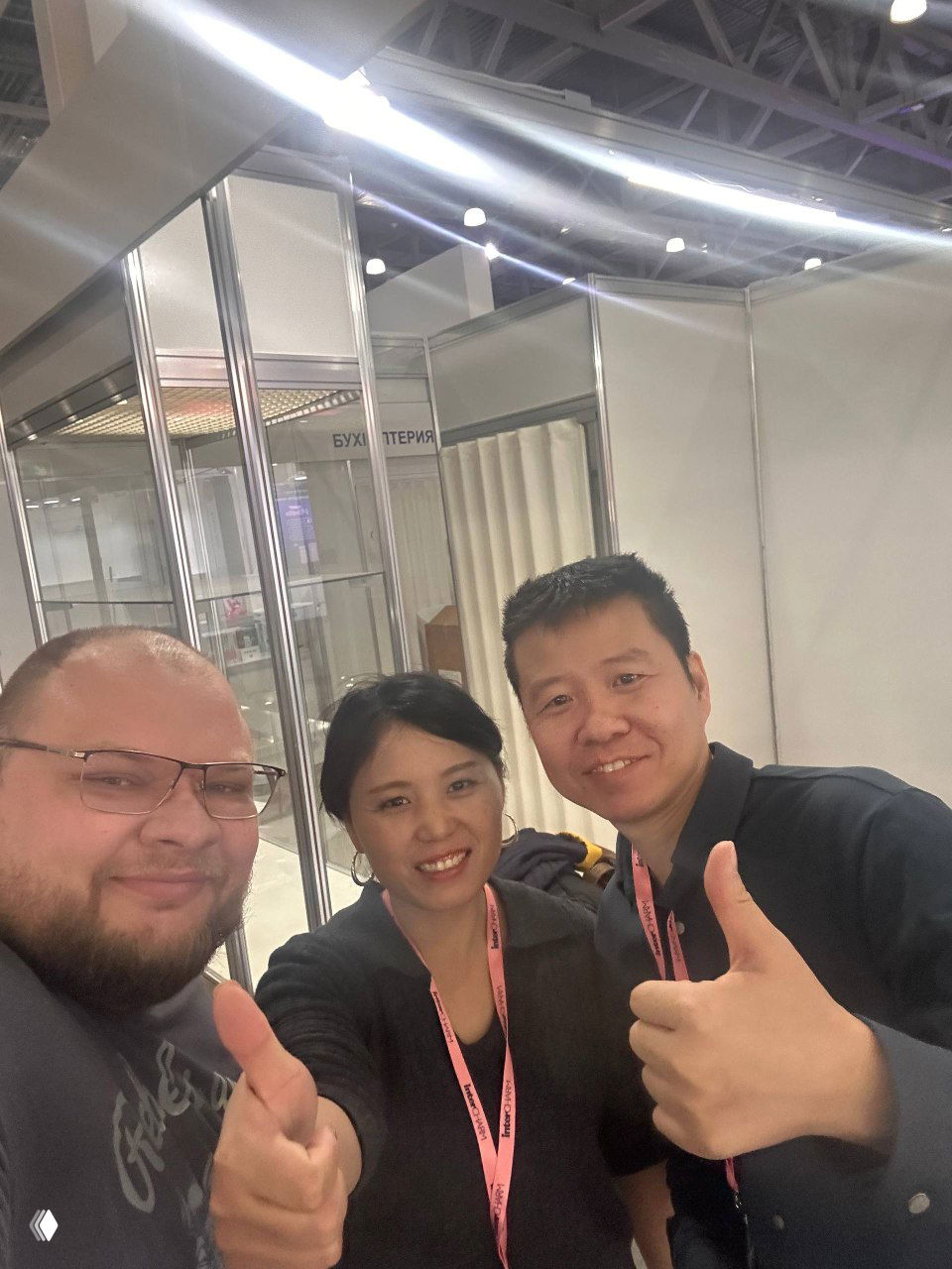Селфи автора с китайскими партнёрами на выставке: трое людей улыбаются и показывают «палец вверх» на фоне выставочного зала.