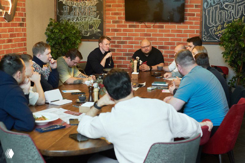 Группа предпринимателей сидит за большим овальным столом во время мастер-класса, на столе бумаги, тетради и бутылки, идёт живое обсуждение.