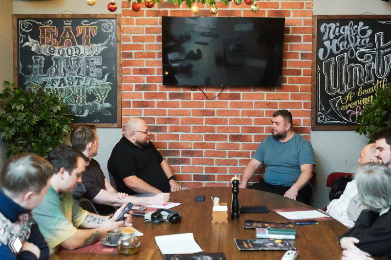 Ведущий беседует с участником напротив, остальные слушают; на столе тетради и фляжки, чувствуется фокус на глубоком психоаналитическом разборе бизнеса.