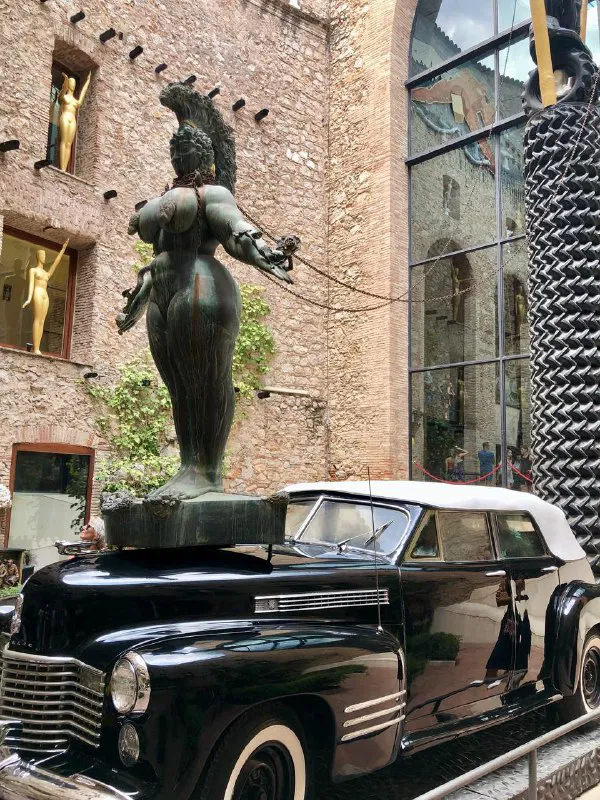 Скульптура в сюрреалистическом стиле и чёрный ретро-автомобиль на открытом дворе — характерная сцена у музея Сальвадора Дали в Фигерасе.