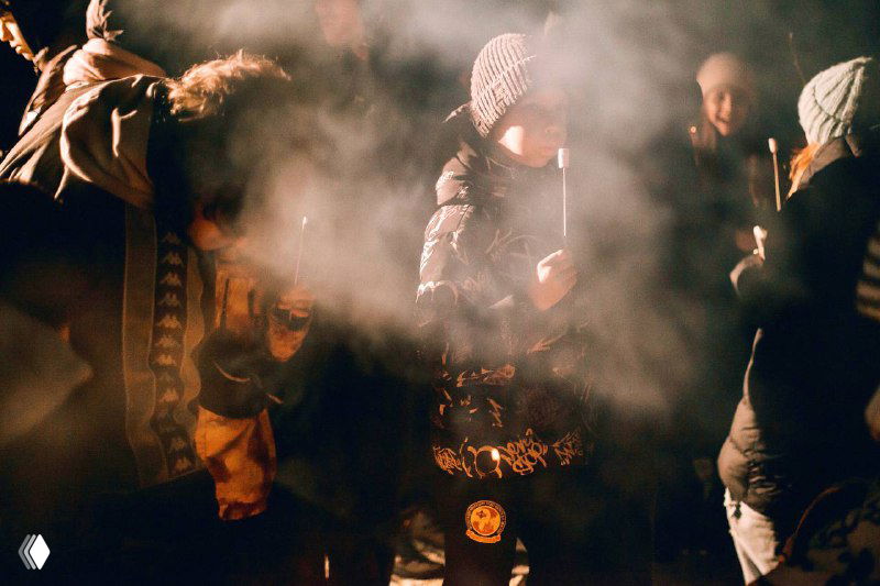 Вечерняя уличная сцена с людьми в тёплой одежде: клубы дыма или пара, приглушённая подсветка и ощущение движения на свежем воздухе.