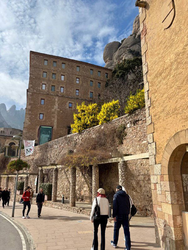 Прогулочная зона у стен монастыря Монсеррат: каменные построения, люди на тропинке и горные вершины на фоне ясного неба в весенний день.