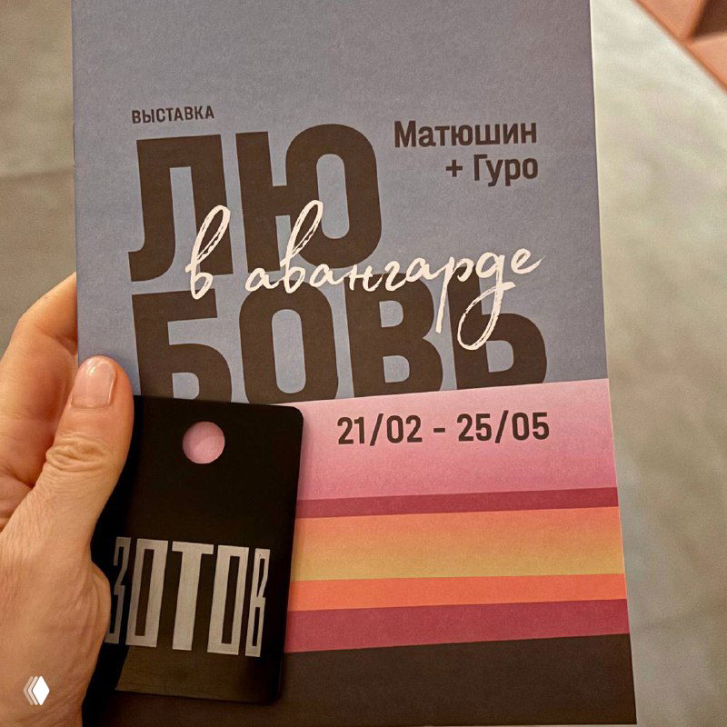 Крупный план буклета и брелока Центра «Зотов» с заголовком выставки «Любовь в авангарде» и датами 21/02–25/05 на фоне пола галереи.