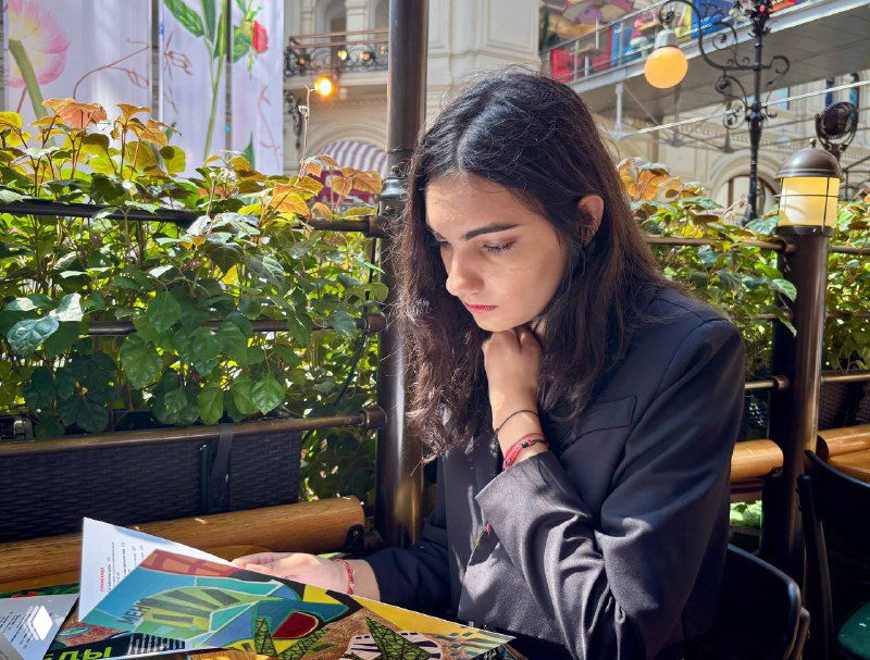 Девушка сидит в кафе с меню на столе у растущих зелёных растений, сосредоточенно рассматривает предложения — атмосфера летнего кафе и городской жизни.