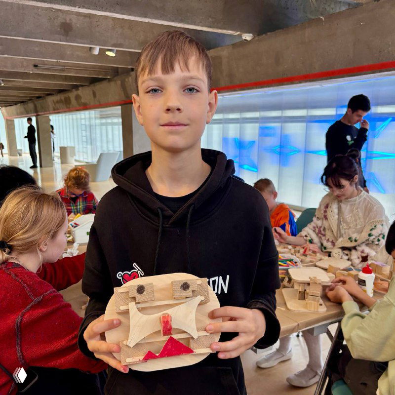 Дети на бесплатном мастер-классе в музее «Гараж», мальчик с деревянной поделкой на фоне других участников и мастерской зоны.