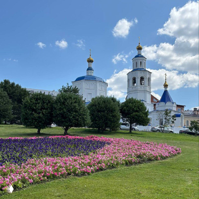 Пышные зелёные газоны и цветник на переднем плане, на заднем плане белое церковное здание с голубыми куполами под ясным летним небом.
