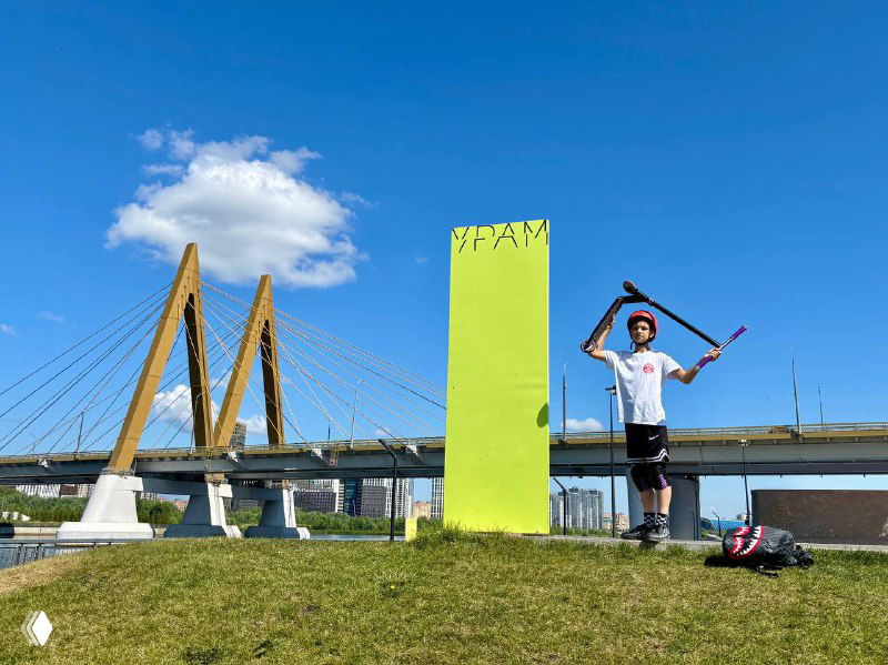 Ребёнок на самокате на травяном холме рядом с ярким зелёным арт-объектом и вантовым мостом на заднем плане; солнечный летний день.