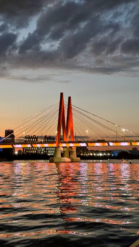 Вечерняя фотография вантового моста, подсвеченного огнями, с отражением в реке; небо в драматических облаках на закате.
