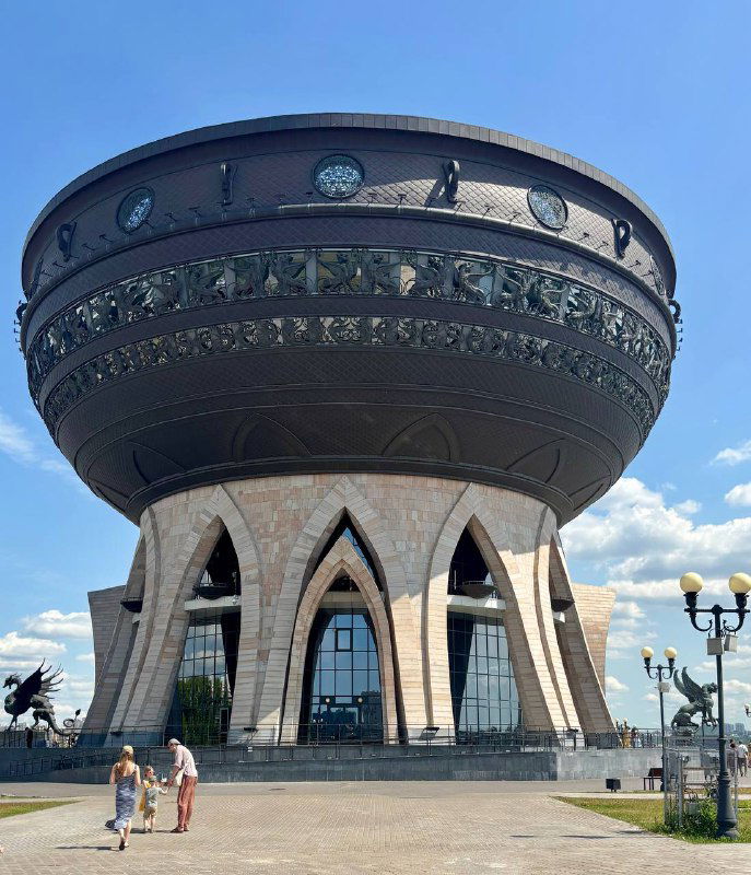 Здание Центра семьи «Казан» в форме огромной чаши с орнаментом по борту, расположенное у набережной и освещённое дневным солнцем.