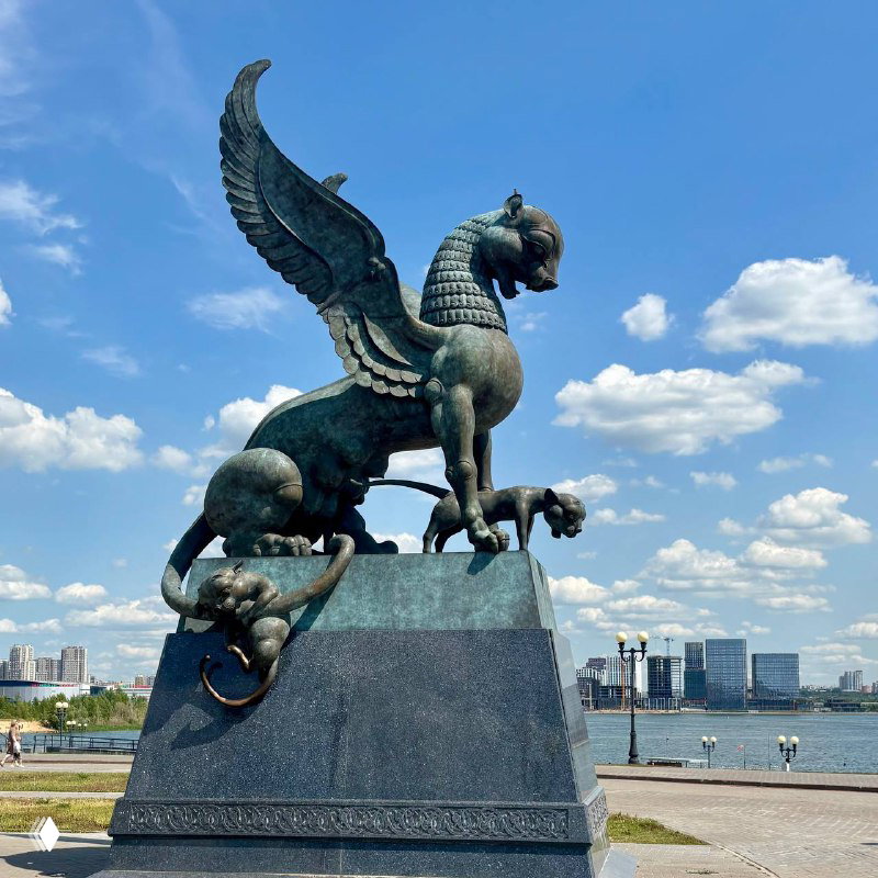 Крупный план бронзовой скульптуры зиланта — крылатого дракона на постаменте, установленного на набережной Казани на фоне неба.