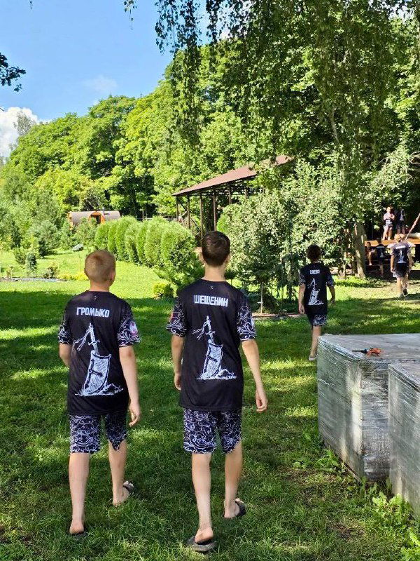 Двое мальчиков в футболках лагеря идут по тропинке к спортивной поляне и беседке, видны элементы лагерной инфраструктуры и другие участники сборов.