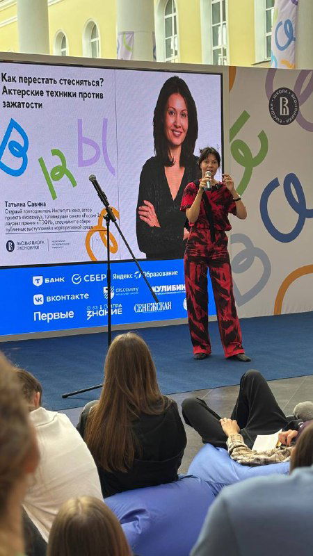 Лектор выступает на сцене перед аудиторией школьников и студентов: большая проекция с фотографией спикера, сидящие слушатели и атмосфера образовательного события в ВШЭ.