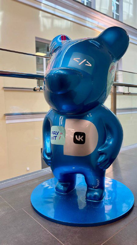Синяя декоративная статуэтка в виде медвежонка с логотипами и стикерами IT/платформ, установленная в зоне фестиваля и служащая элементом оформления и фотозоны мероприятия.