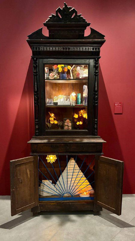 Антикварный шкаф с открытыми дверцами и внутренней витриной, где выставлены флаконы и предметы быта — часть исторической инсталляции.