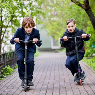 Два мальчика на самокатах катаются по дорожке у школьного здания — активные перемены и детская мобильность на территории образовательного учреждения.