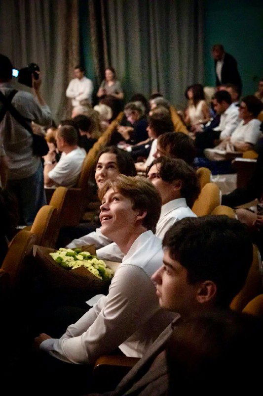 Зрители в актовом зале школы во время школьного мероприятия — родители и ученики смотрят выступление, тёплая атмосфера школьных праздников и собраний.