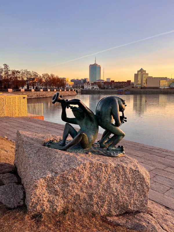 Скульптура у набережной Челябинска на фоне воды и городской застройки при тёплом вечернем небе и спокойной воде.