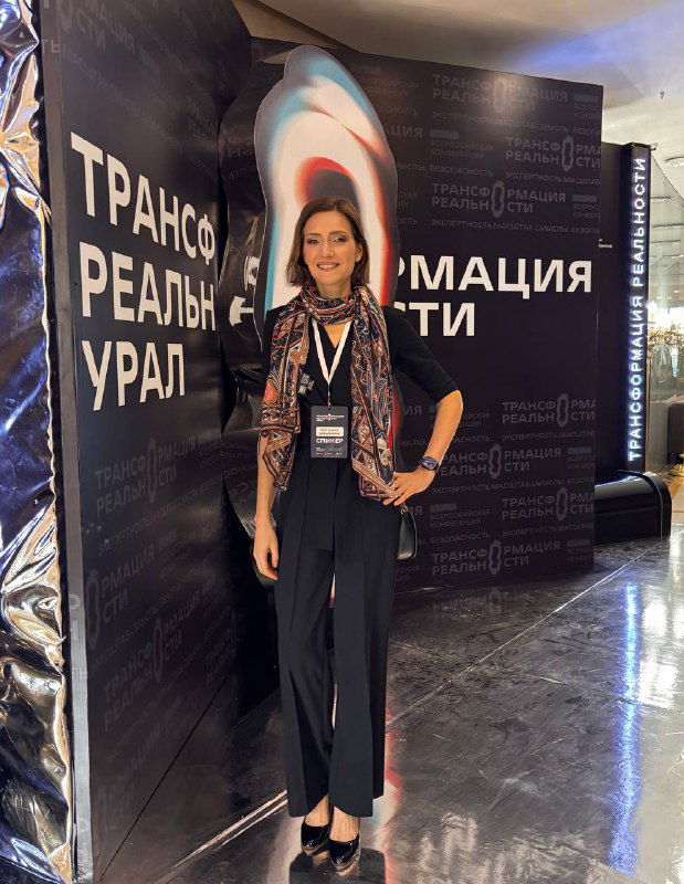 Виктория стоит у бренд‑волла форума «Трансформация реальности. Урал», на ней бейдж спикера, видна сцена и логотип мероприятия.