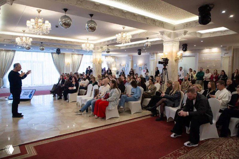 Широкий кадр зала с аудиториями и выступающим спикером на сцене: участники форума сидят и слушают презентацию.