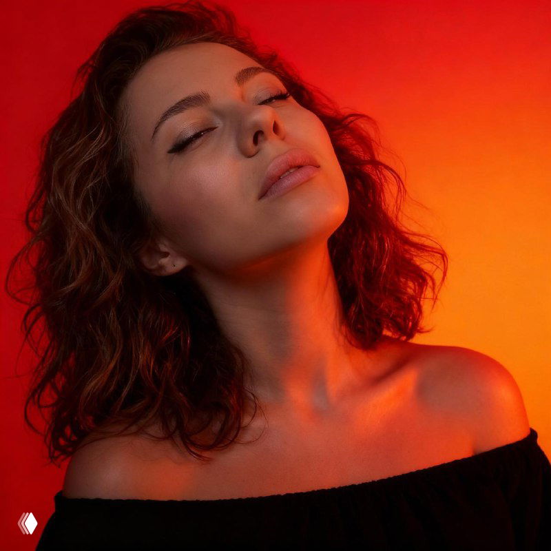 Крупный план портрета женщины с полузакрытыми глазами, сияющая кожа и контрастное красно‑оранжевое студийное освещение на градиентном фоне.