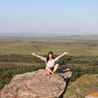 Женщина сидит на камне на вершине холма с расправленными руками, пейзаж степи на заднем плане — символ достижения и свободы после смены профессии.