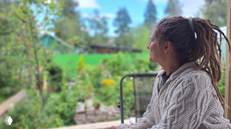 Женщина в свитере с дредами смотрит в сторону сада: вид с террасы, расслабленное состояние и природный фон с зеленью и огородом.