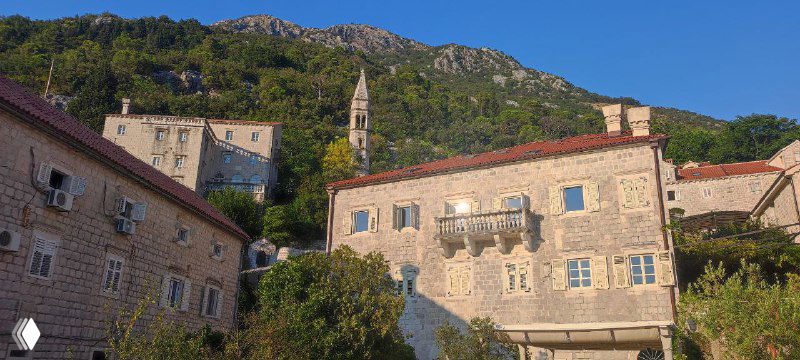 Каменные дома Пераста на склоне холма под голубым небом: старинная архитектура, зелёные насаждения и церковная колокольня вдалеке.