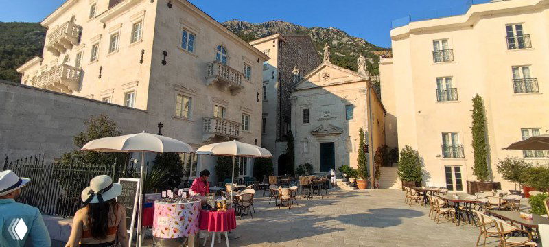 Площадка с уличными столиками и зонтиками в Перасте: туристы, торговая палатка и атмосфера небольшой прибрежной жизни на закате.