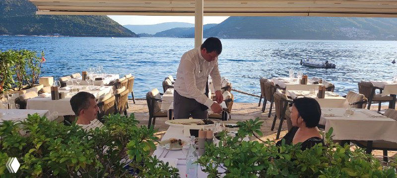 Рестораны на набережной Пераста: официант сервирует столики у самой воды, виднеются горы и спокойная морская гладь вечером.