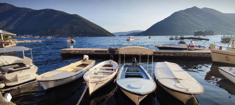 Небольшие моторные лодки пришвартованы в порту Пераста, вид на водную гладь и отдалённые горные силуэты под ясным небом.