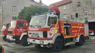 Пожарная машина на городской площади Пераста: несколько машин в ряду и снаряжённый пожарный в красной форме, вечерняя сцена.