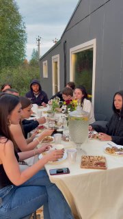 Гости за длинным столом на ретрите: участники едят и беседуют, на столе цветы и блюда — сцена совместного питания и общения в группе