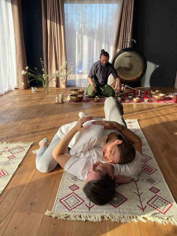 Ведущий и участник лежат на матах в зале рядом с гонгом и поющими чашами — момент телесной практики и доверительного контакта внутри группы