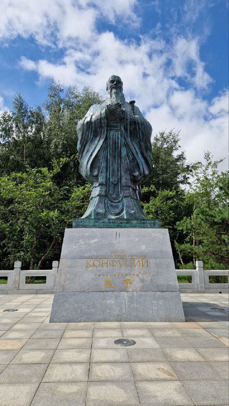 Бронзовый памятник Конфуцию на каменной тумбе на фоне неба и зелёных деревьев в парке «Хуамин», вид с уровня посетителя.