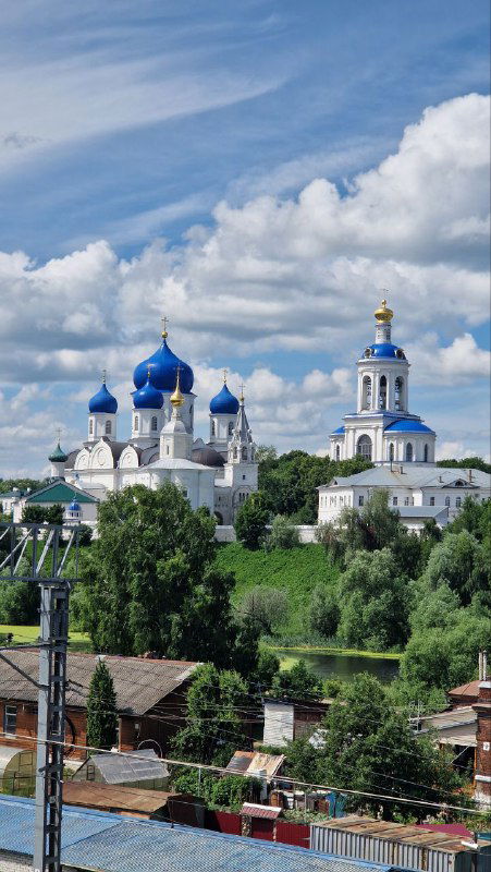 Вид на монастырский ансамбль с синими куполами и золотыми главками в Боголюбово: крыши домов внизу и панорама на холме под ярким небом.
