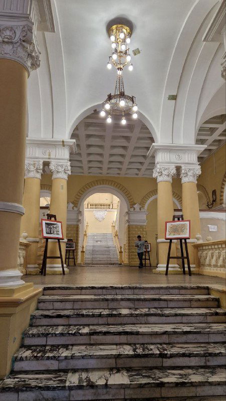 Вестибюль и лестница внутри здания с мраморными ступенями, арками и люстрой — вид снизу на главный вход и экспозиционные таблички.