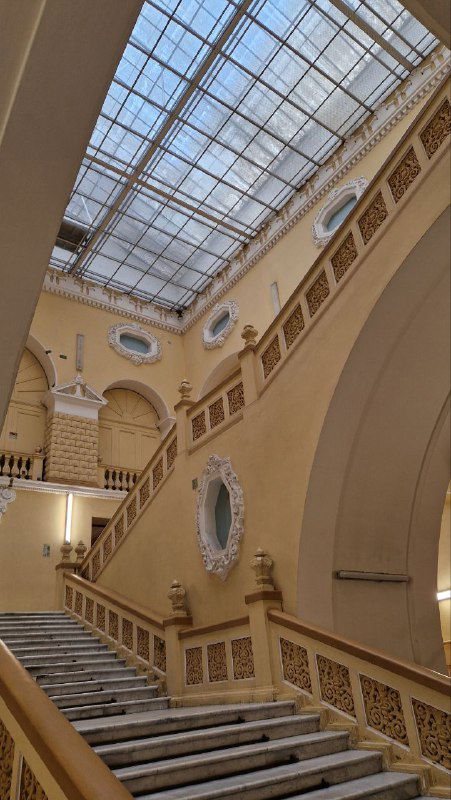 Большая лестничная площадка под стеклянным потолком-фонарём; световой купол и балконы второго уровня в интерьере здания.